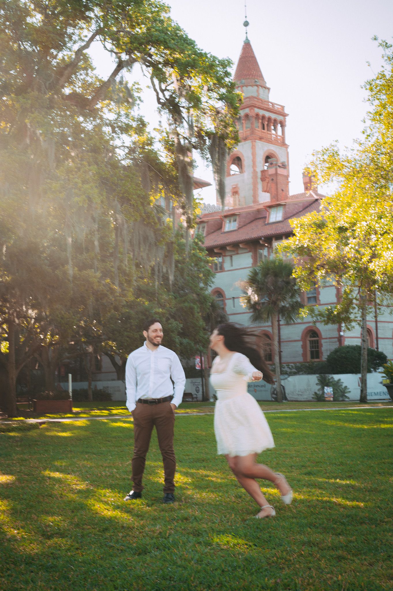 Couple dancing on green lawn with historic red brick tower building in golden sunlight background.