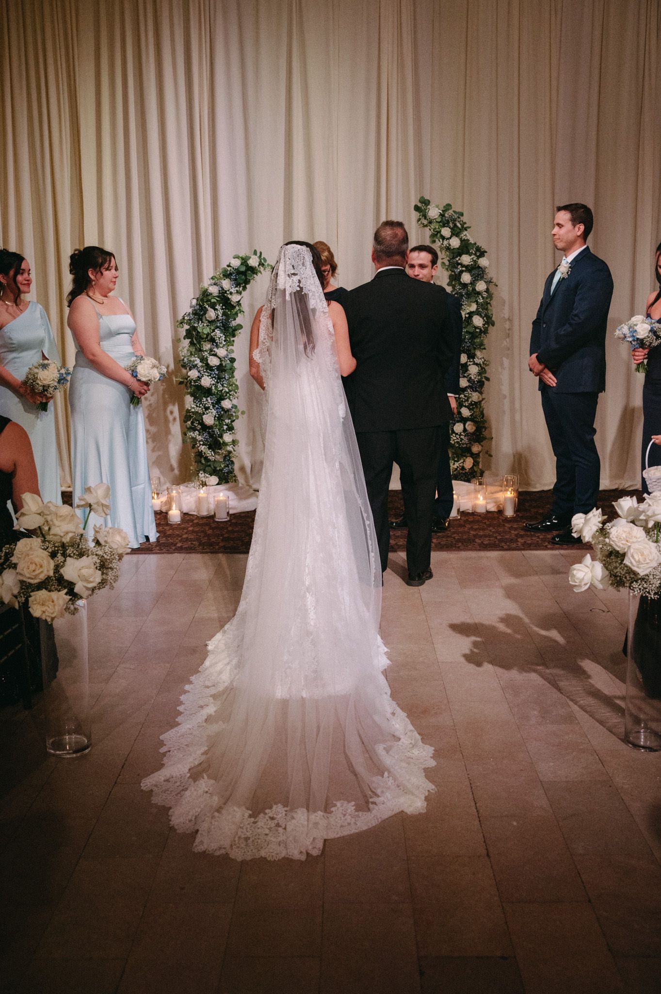 Bride in lace cathedral veil walks toward floral arch altar during elegant indoor wedding ceremony.