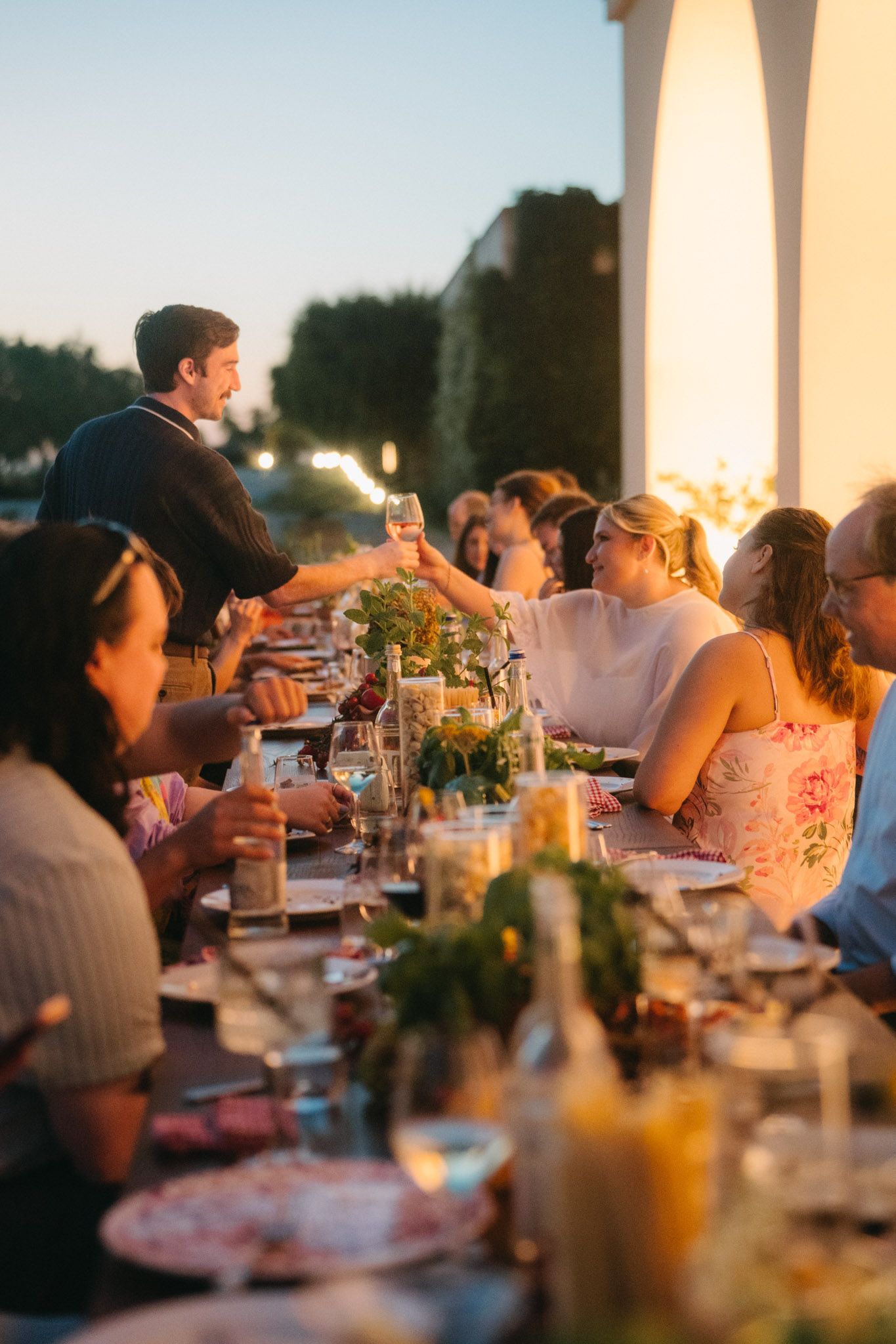 Guests toasting at an elegant outdoor dinner party at dusk, with a long table set with flowers and candles.