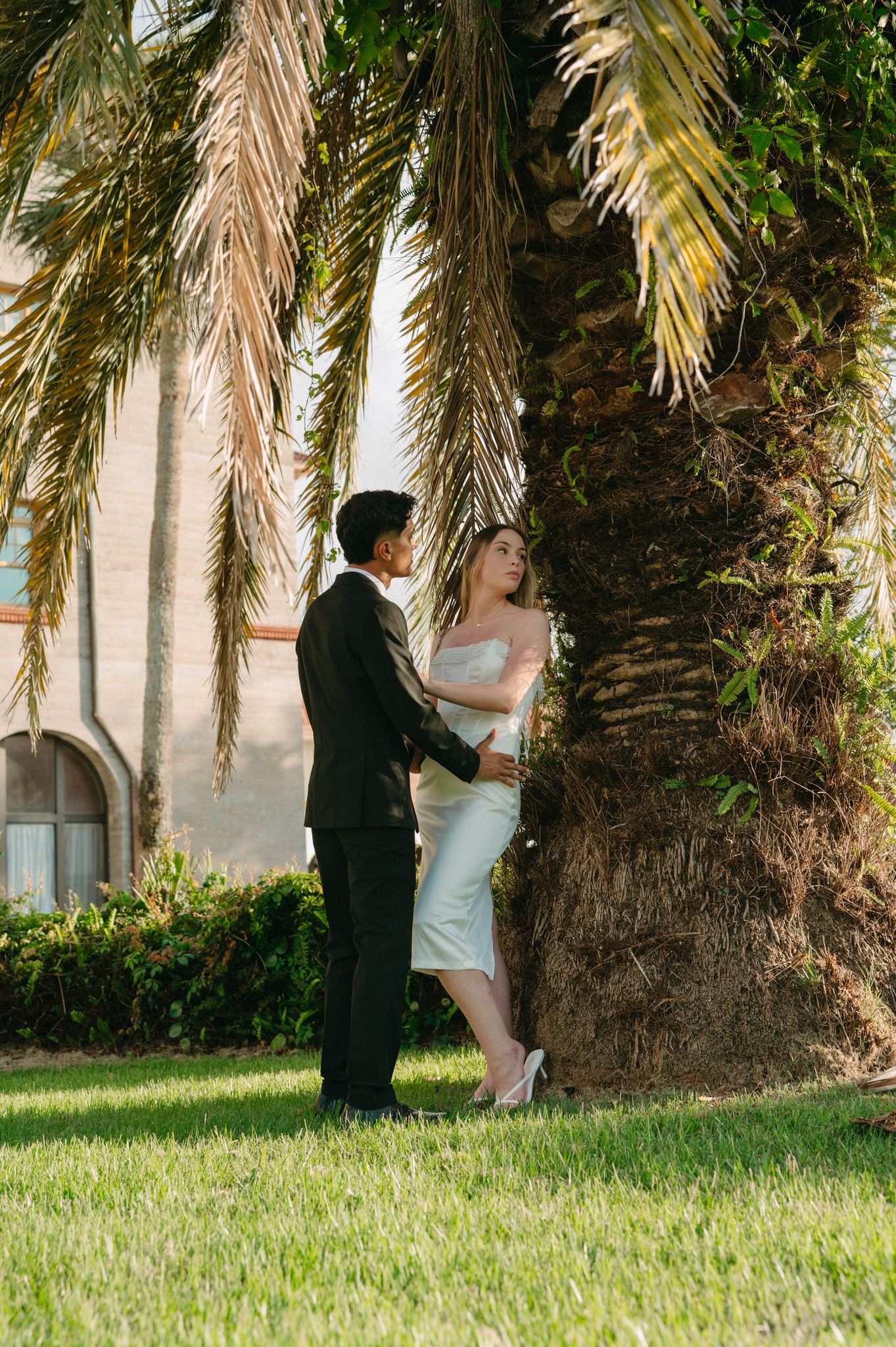 Bride and groom sharing intimate moment under tropical palm tree on lush green lawn.