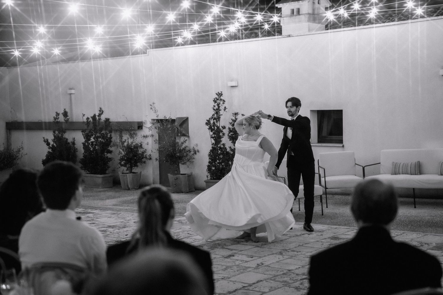 Black and white photo of a bride and groom sharing their first dance outdoors under string lights at a wedding reception.