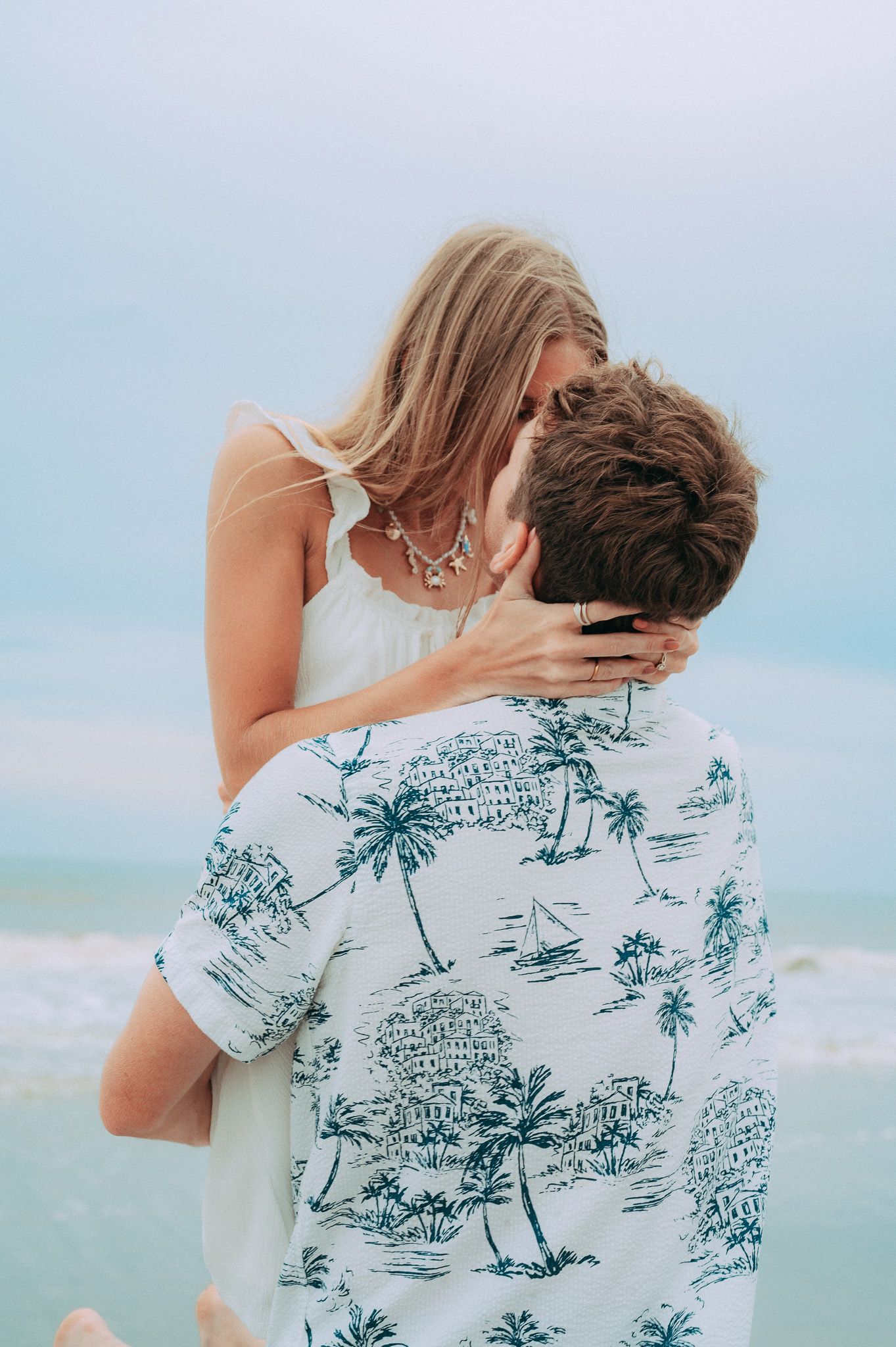 Couple sharing a romantic kiss on the beach, man in palm tree shirt lifting woman in white dress by the ocean.