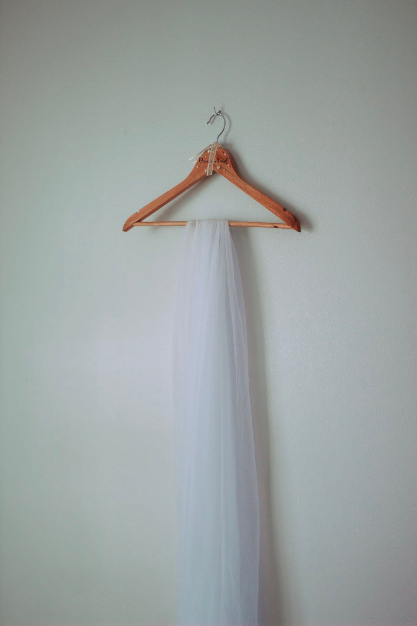 Minimalist white wedding veil hanging on wooden hanger against pale green wall.