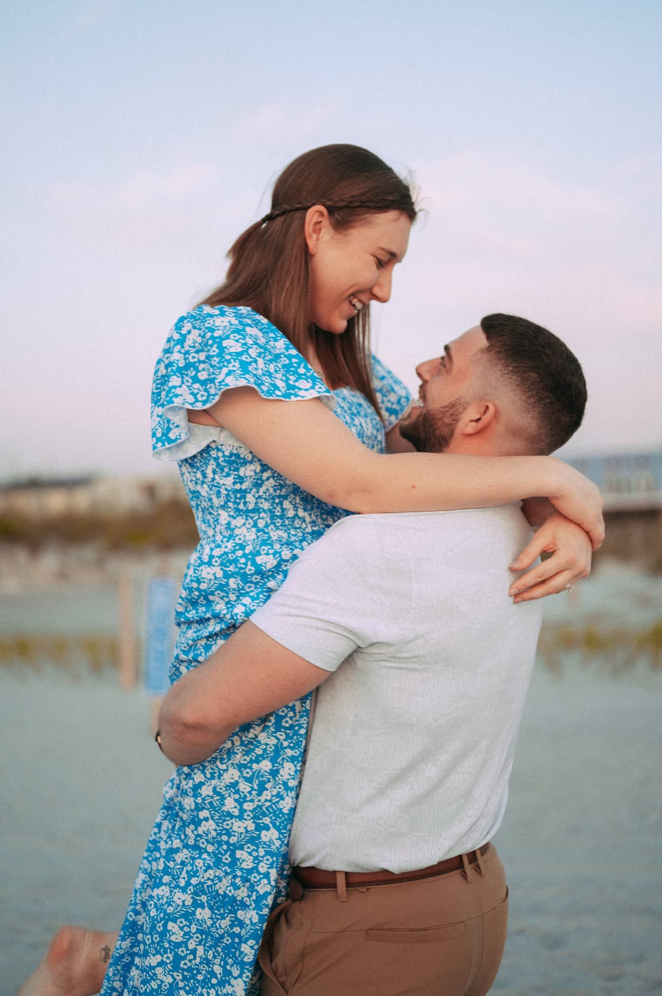 Happy couple embracing on a beach at sunset, woman in blue floral dress lifted by man.