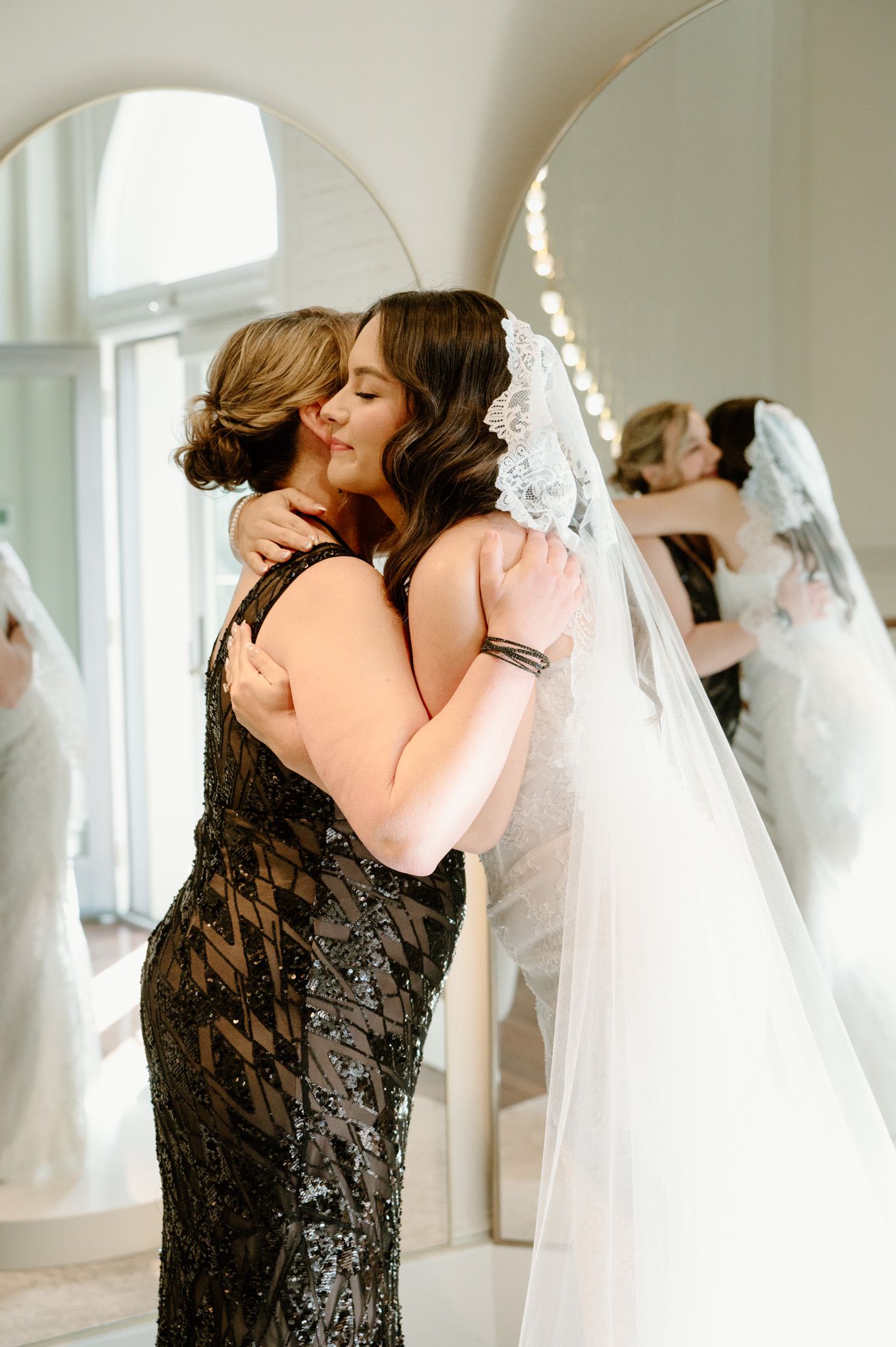 Bride in white gown and veil shares emotional hug with guest in black beaded dress near elegant mirror.