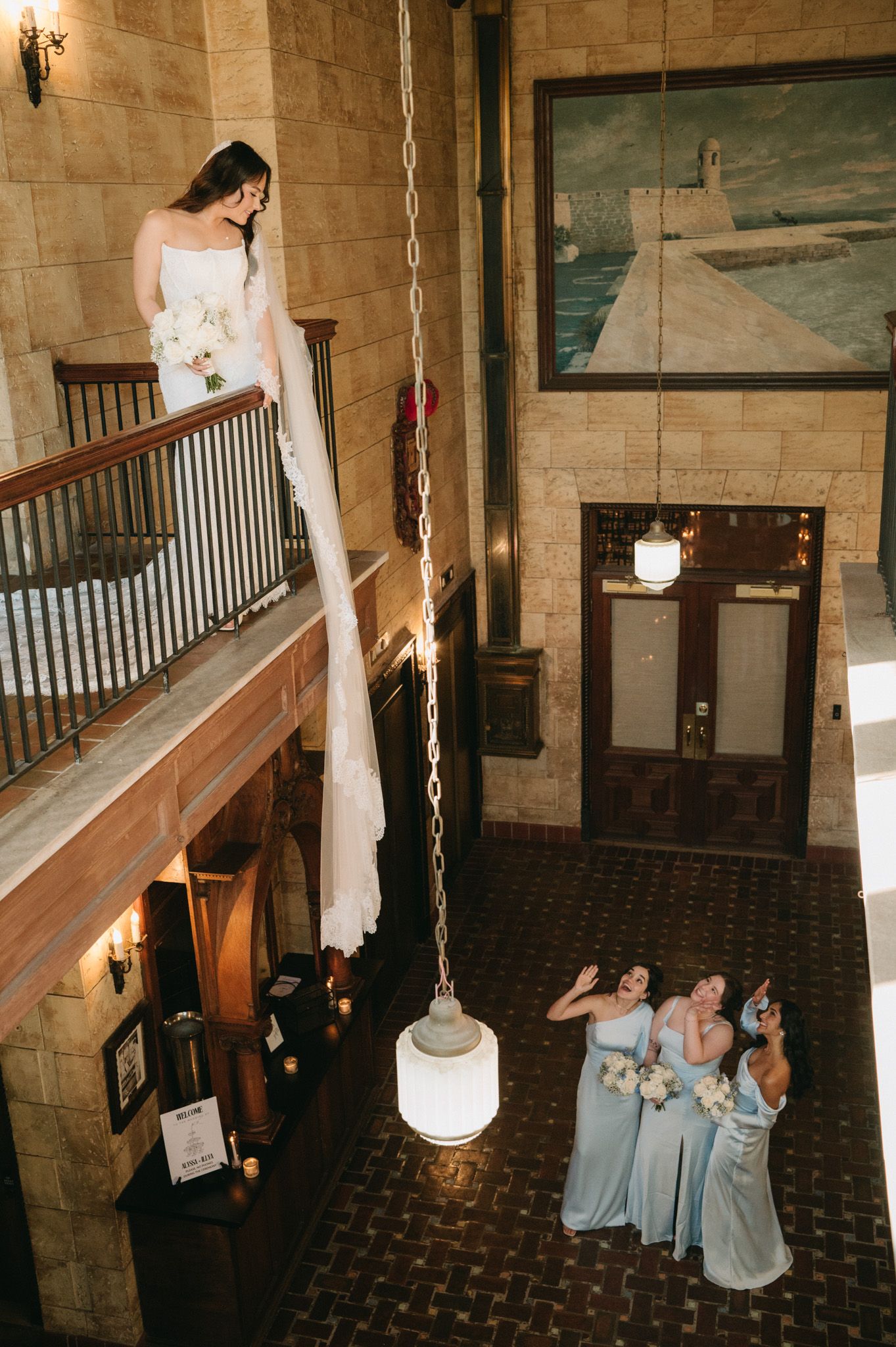 Bride in white gown stands on balcony above bridesmaids in blue dresses in elegant stone venue.