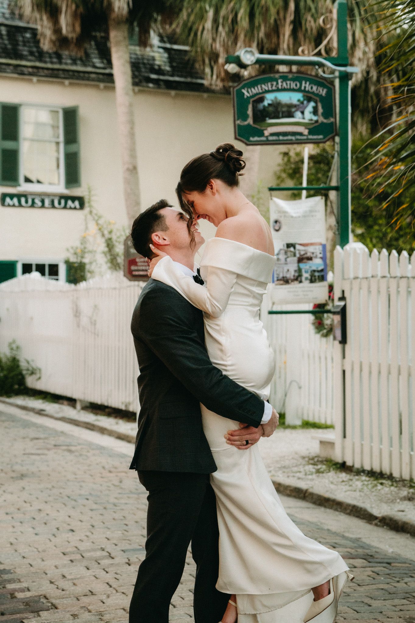 Groom lifts bride in off-shoulder white gown for a kiss on a cobblestone street near a historic museum.
