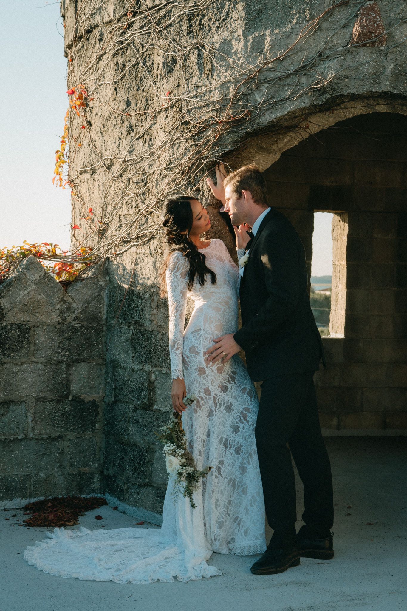 Bride in long-sleeve lace gown and groom kiss beneath a stone archway draped with autumn vines at golden hour.