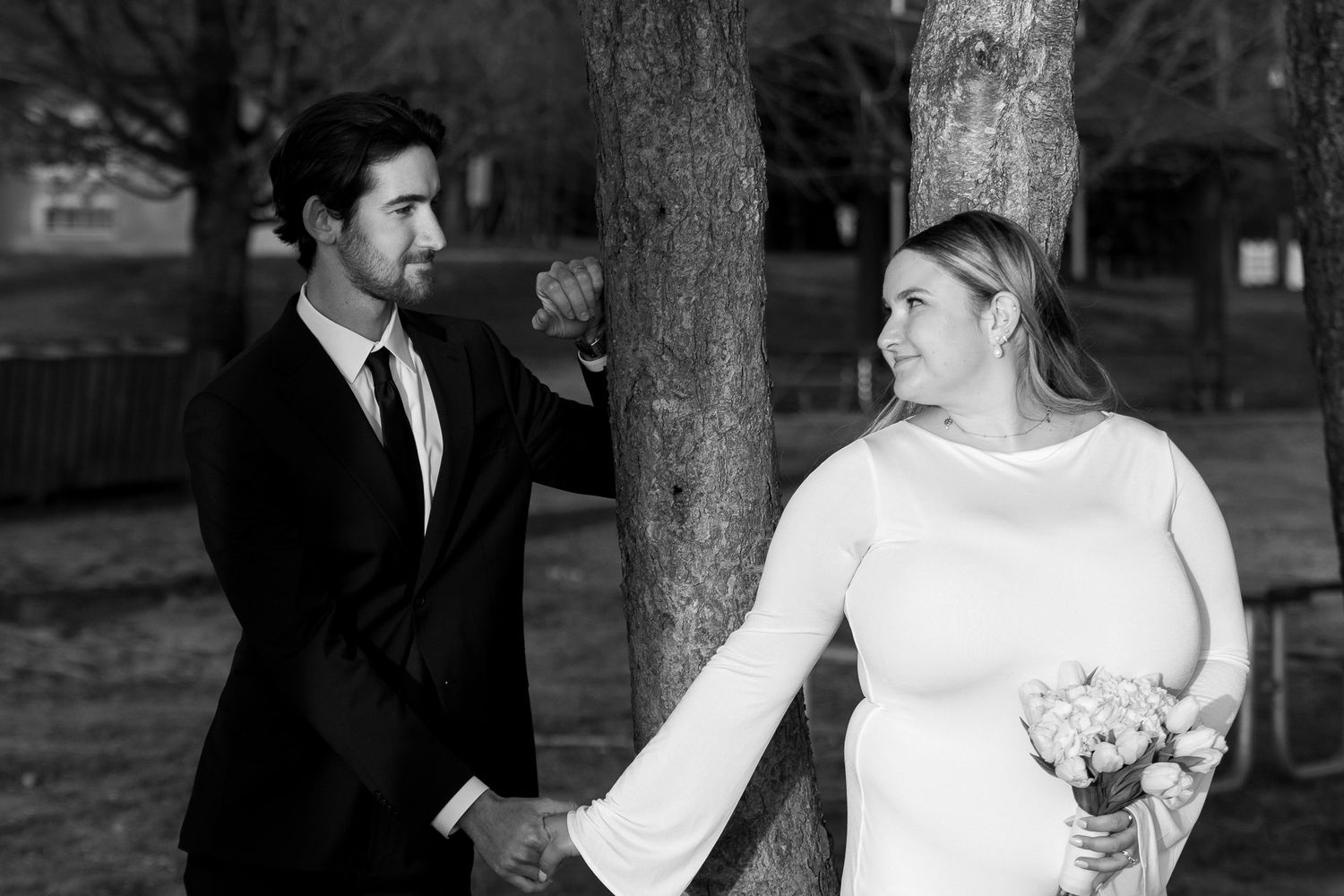 Black and white photo of newlyweds holding hands beside a tree in a park.