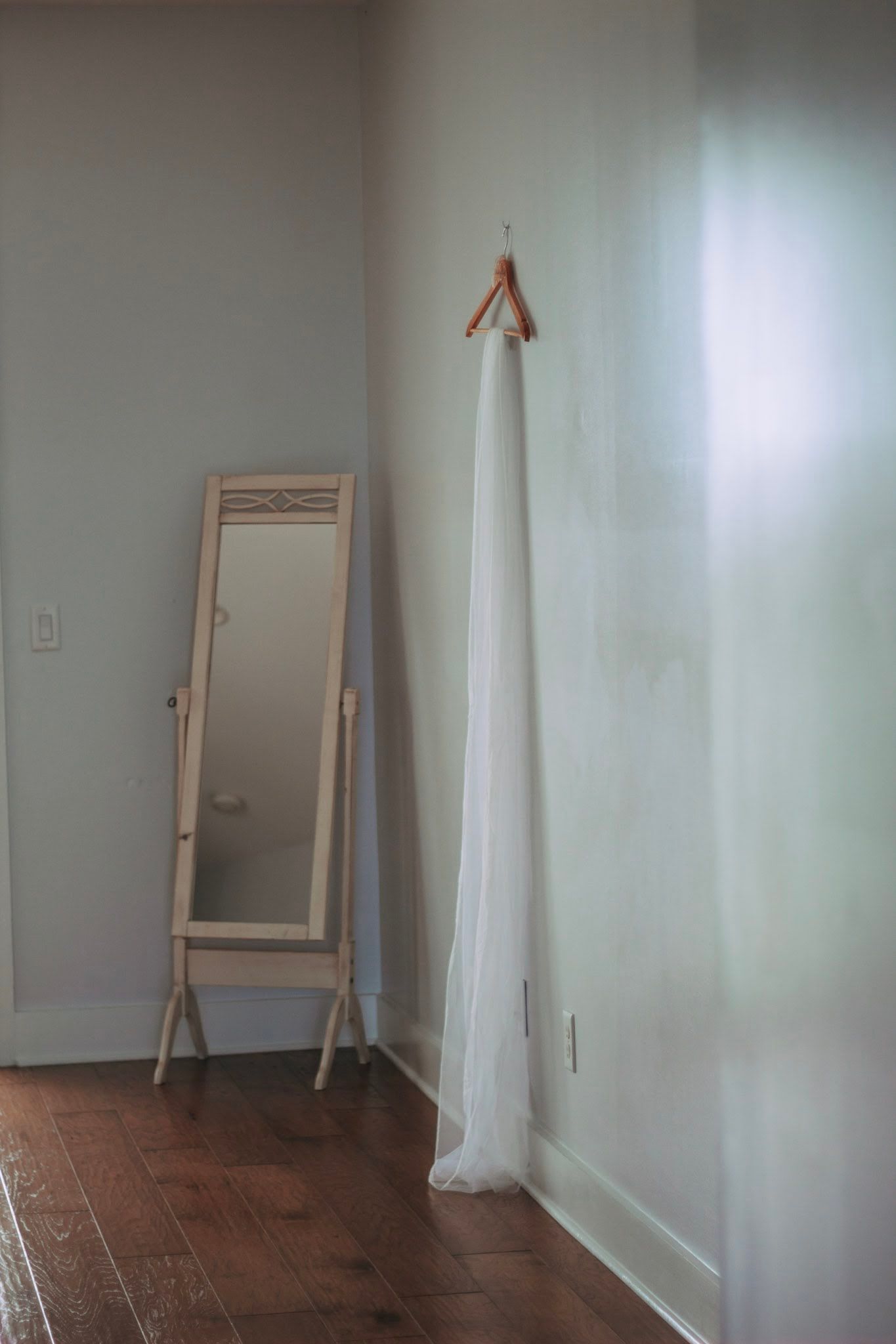 White wedding dress on wooden hanger beside vintage full-length mirror on hardwood floor.