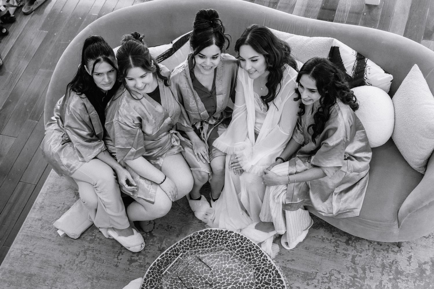 Bride and bridesmaids in robes huddle together on a curved sofa, captured in black and white from above.