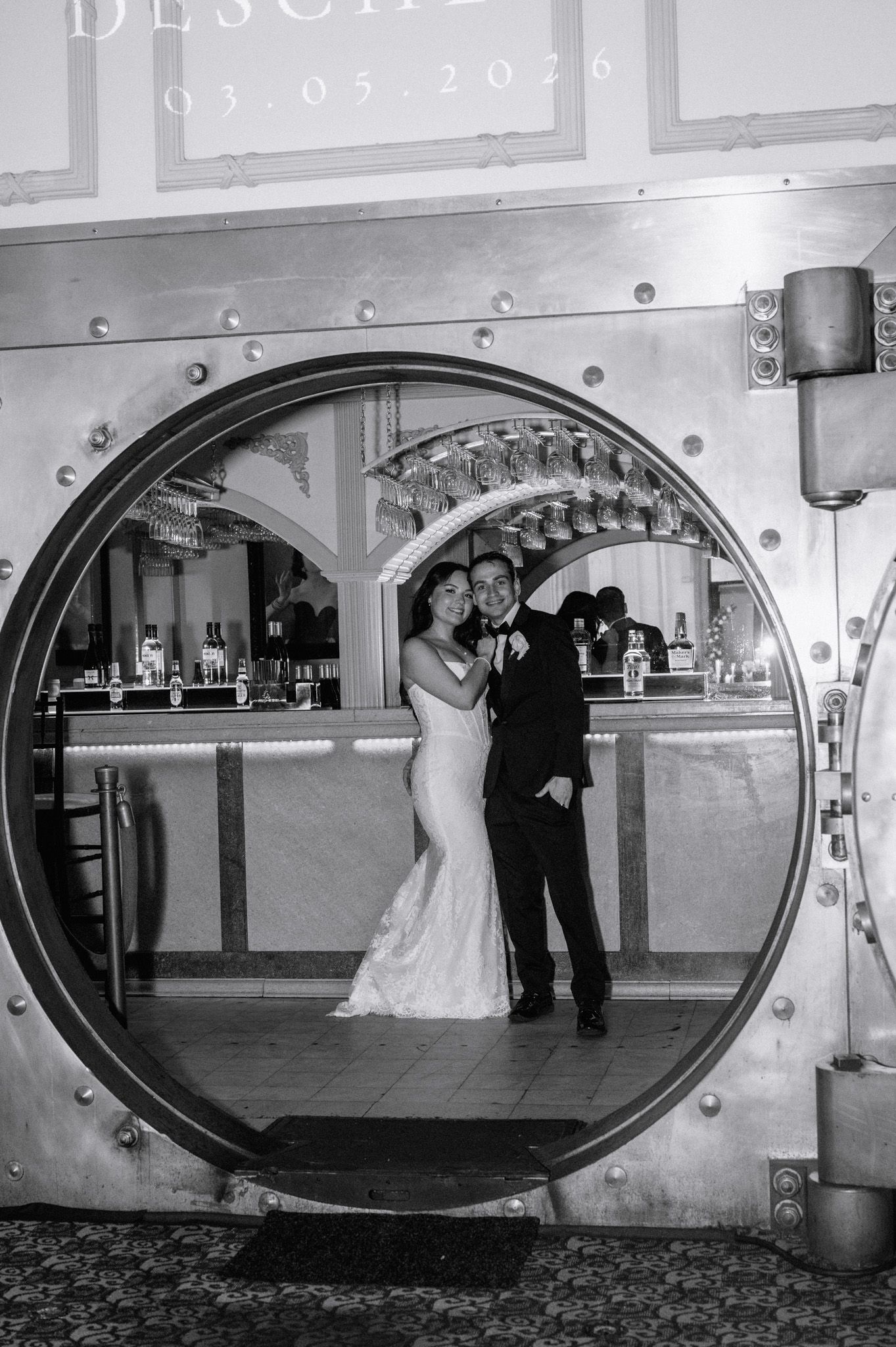 Black and white wedding photo of bride and groom posing inside a large vault door at an elegant venue.