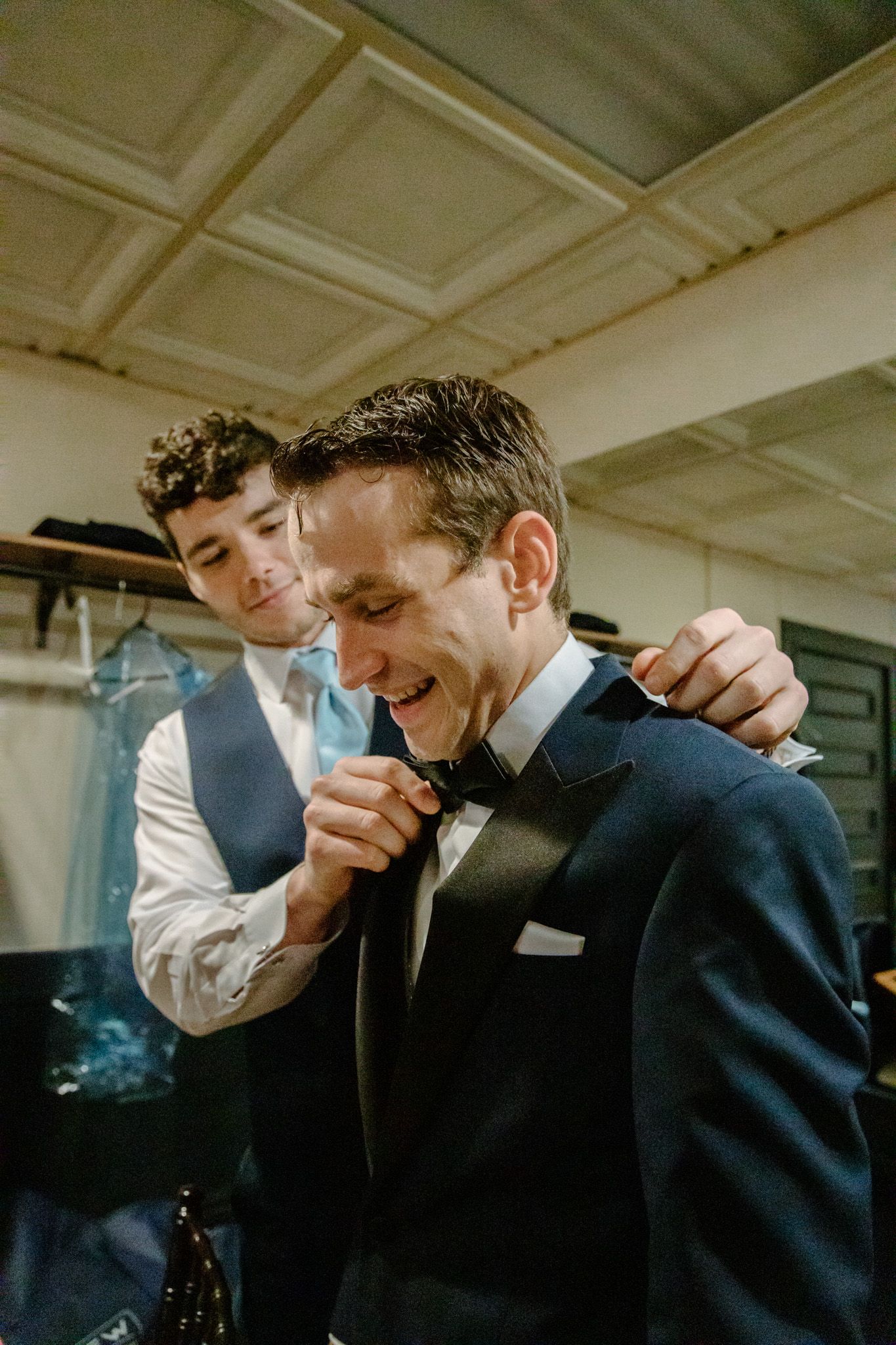 Groom smiling as groomsman helps adjust bow tie while getting ready in a dressing room before wedding ceremony.