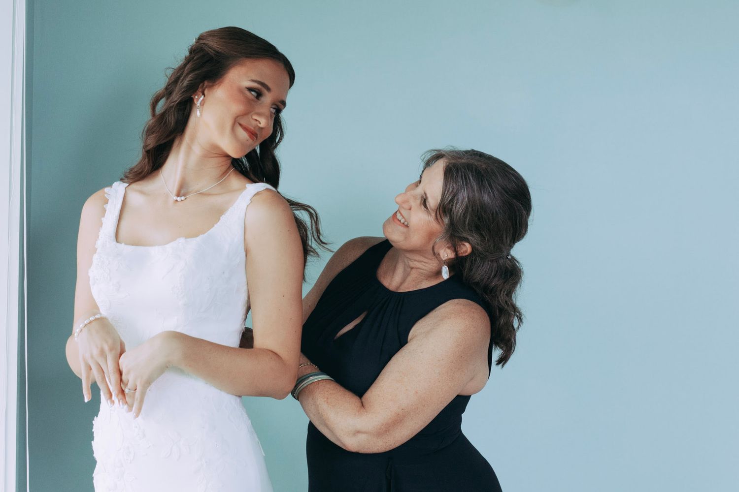 Bride in white dress smiling as mother in black dress lovingly adjusts her gown.
