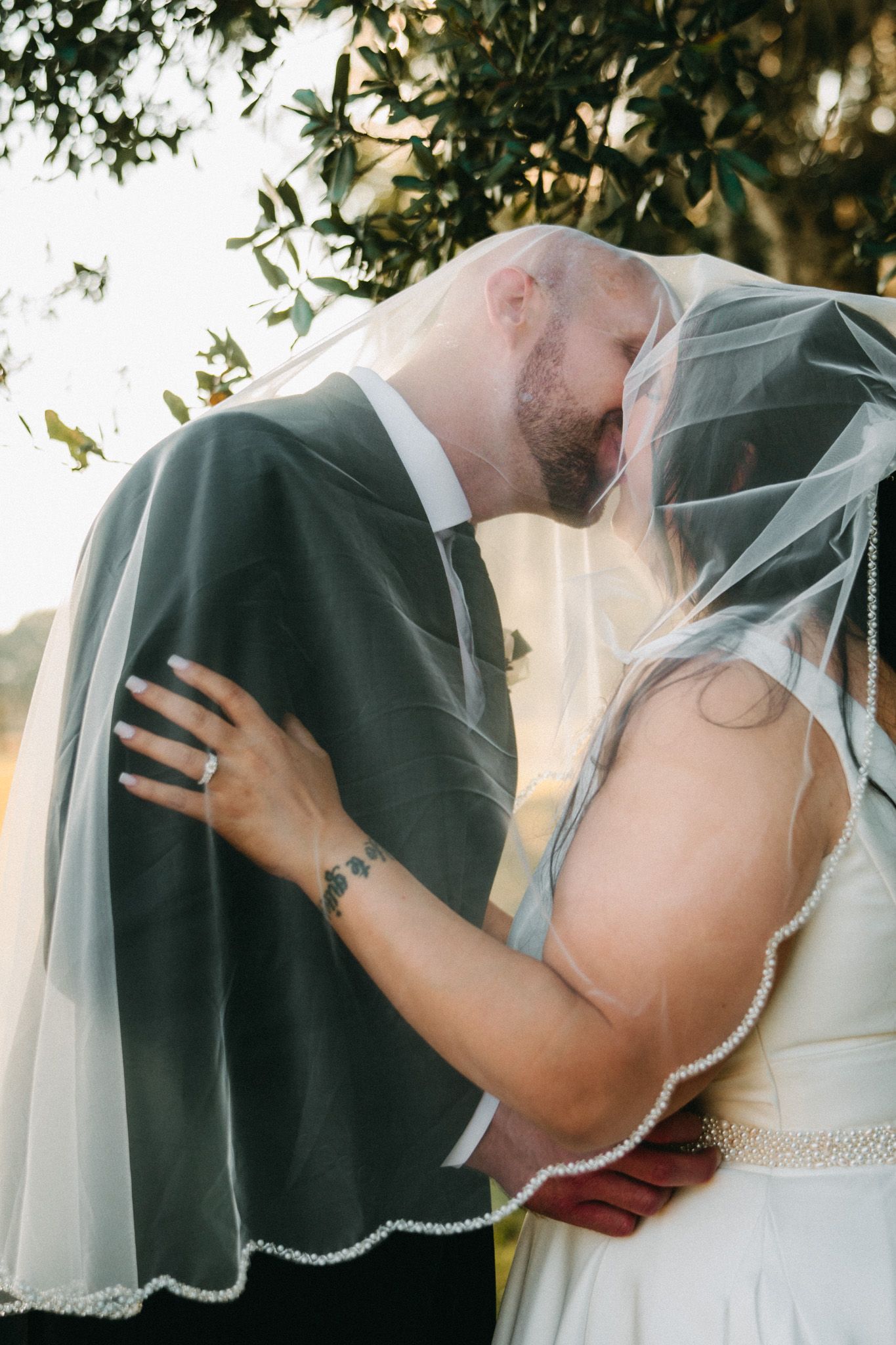Bride and groom share romantic kiss under flowing veil with golden backlight filtering through trees.