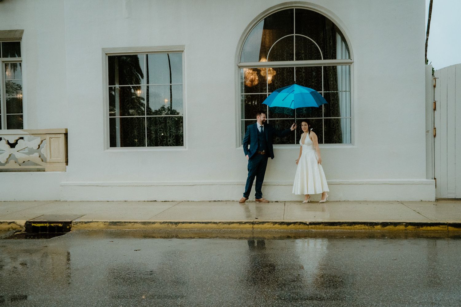 Bride and groom standing apart in rain, bride holding blue umbrella outside white building.