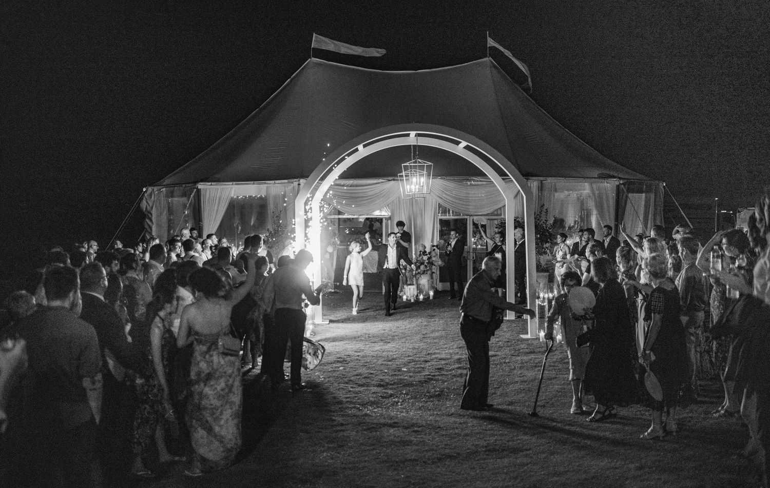 Black and white photo of wedding guests gathered under an illuminated tent entrance at night.