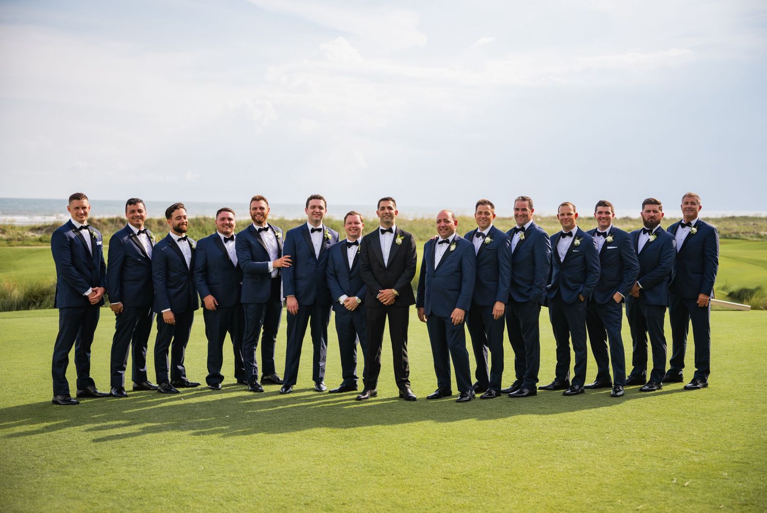 A large group of groomsmen in navy suits stand together on a green golf course for a wedding photo.
