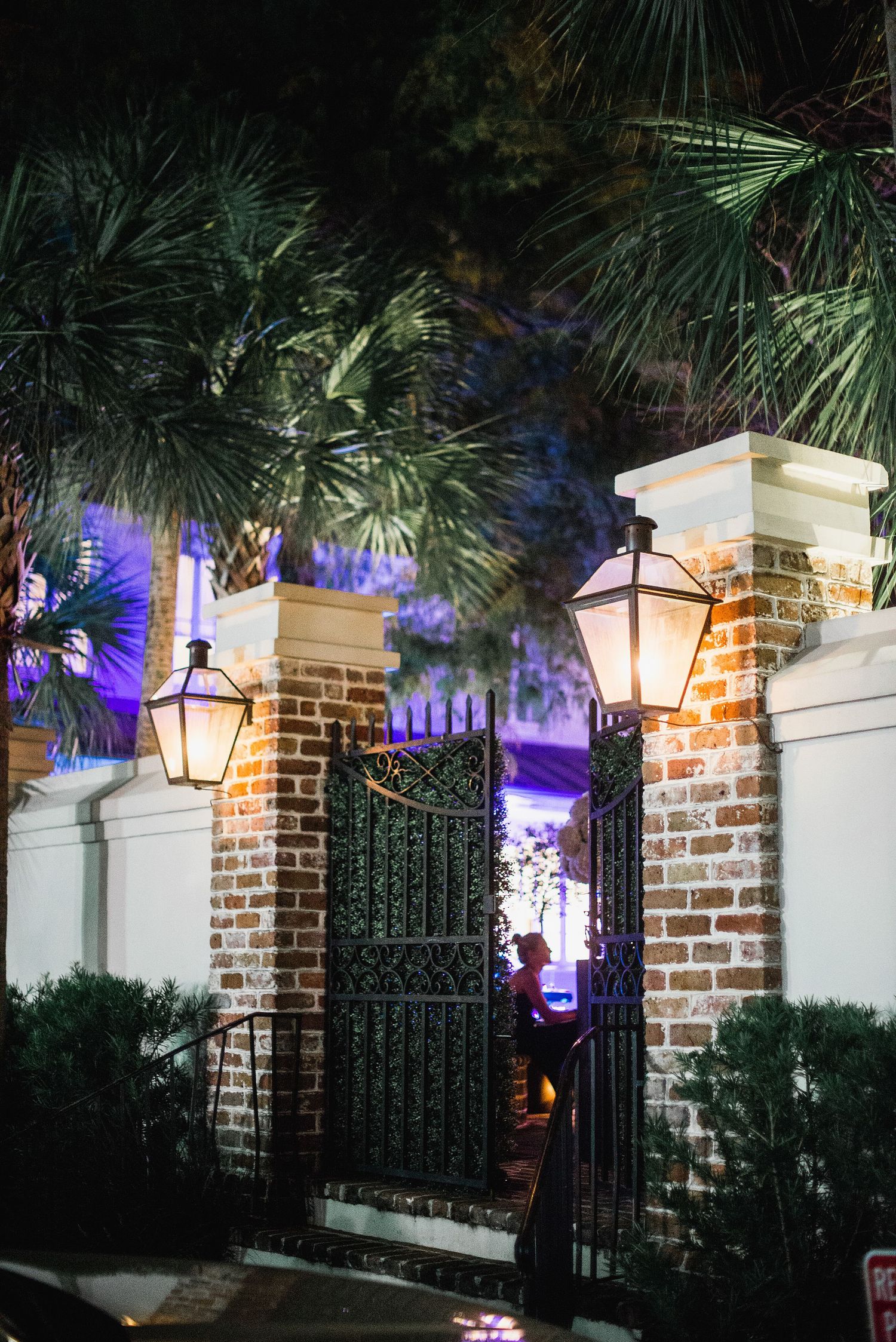 Decorative gas lanterns illuminate a brick and iron gate entrance with palm trees at night, casting a purple glow.