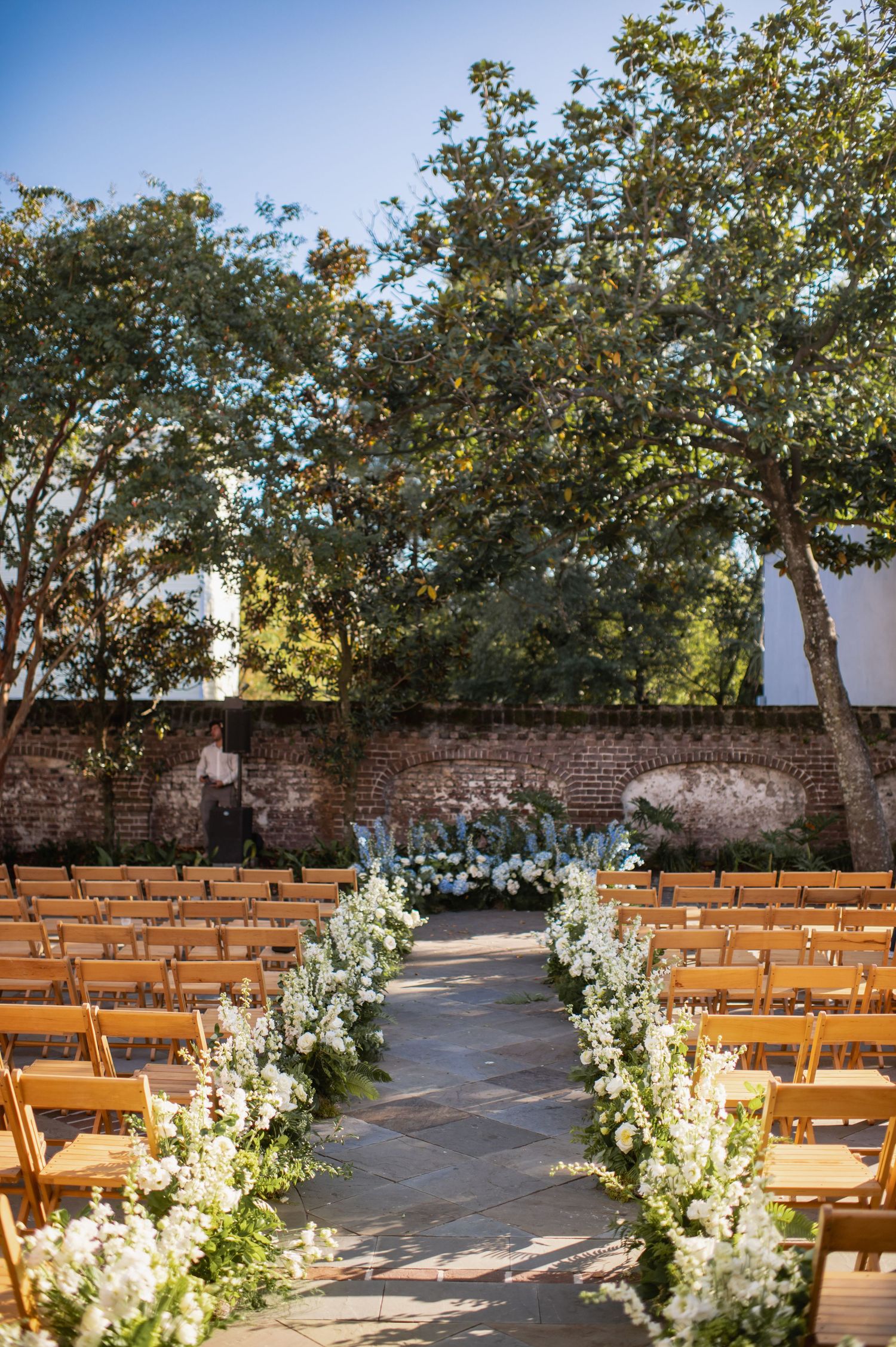 Outdoor wedding ceremony setup with wooden chairs and white floral aisle decorations against a rustic brick wall.