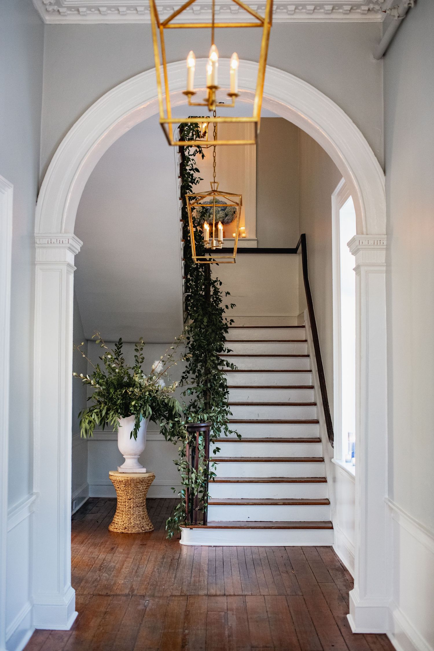 Elegant arched entryway with white trim leads to wooden staircase, featuring yellow lantern and climbing ivy decor.
