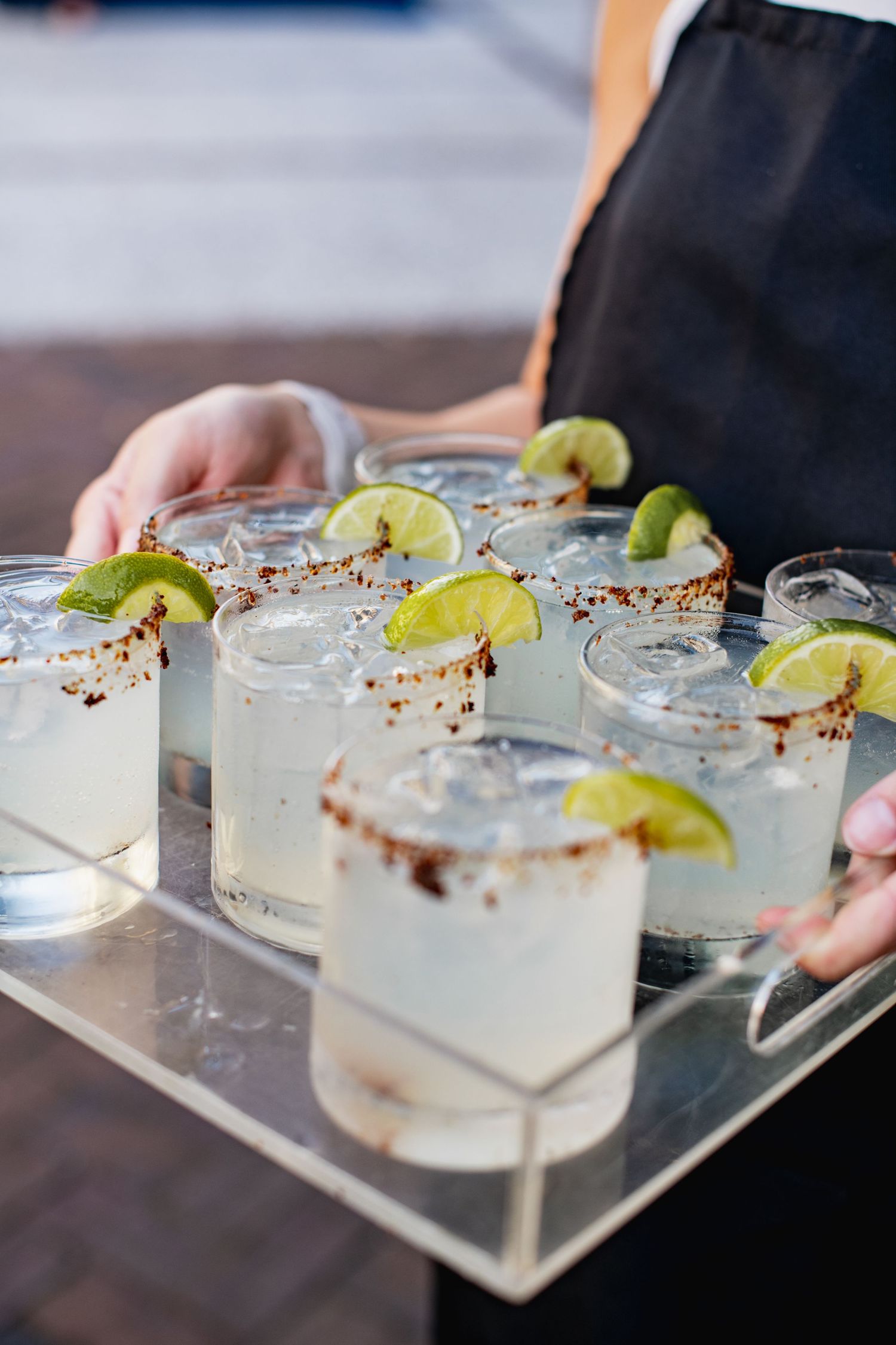 Several margarita cocktails with lime wedges and spiced rims are being served on a clear glass serving tray.