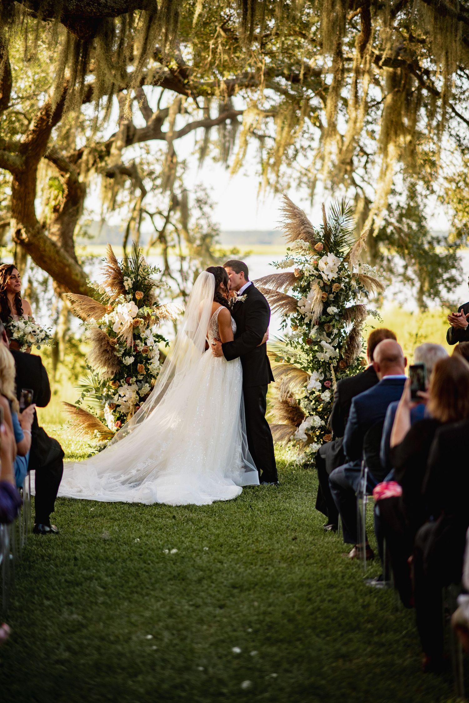 Wedding ceremony under Spanish moss draped oak trees with guests gathered on a grassy lawn at sunset.