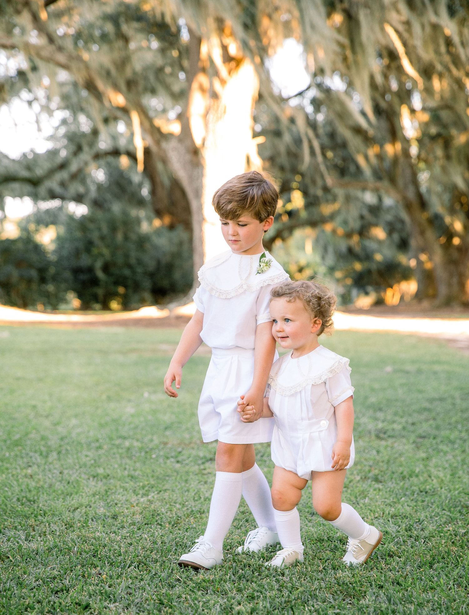 Two children in white outfits and knee socks walk together on grass under Spanish moss-draped trees at sunset.