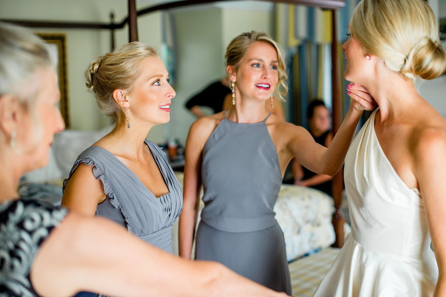 Bridesmaids in matching gray dresses help the bride get ready in a sunlit room before the wedding ceremony.