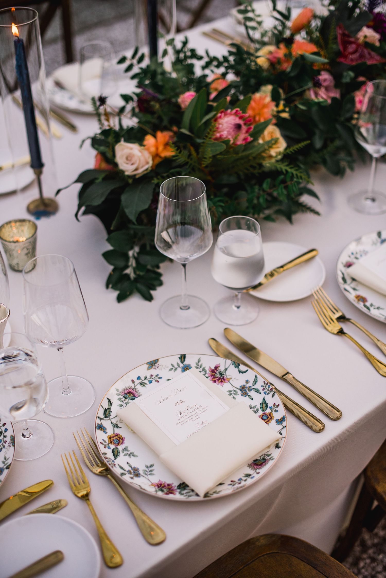 Elegant table setting with floral plates, gold cutlery, wine glasses and coral flower centerpiece on white tablecloth.