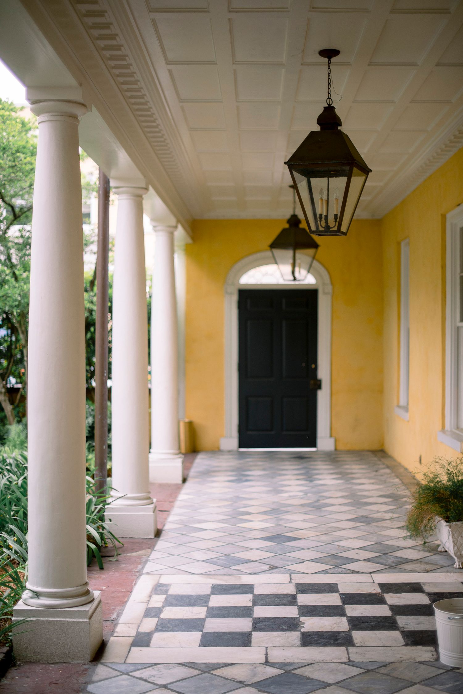 Elegant colonial corridor with yellow walls, white columns, and black and white checkered marble floors illuminated by lanterns.
