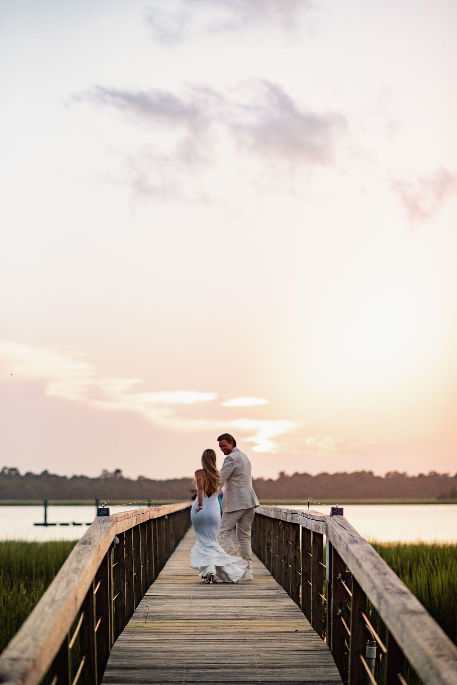A couple stands together on a wooden dock at Lowndes Grove overlooking marsh waters during a dreamy sunset.