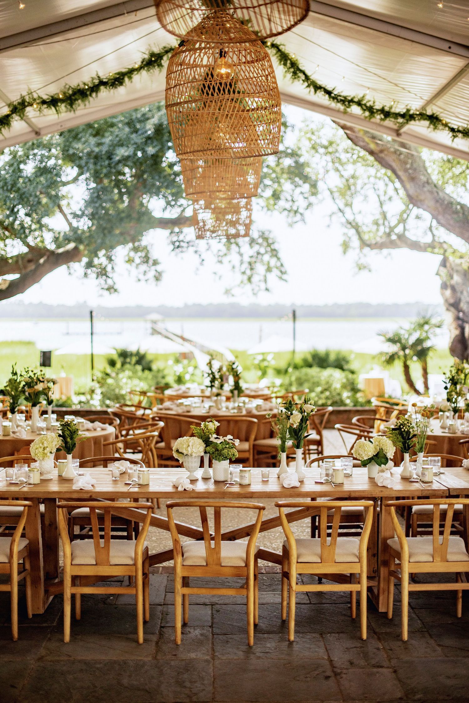 Long rustic wooden tables set for an elegant outdoor wedding reception under a tent with woven rattan chandeliers.