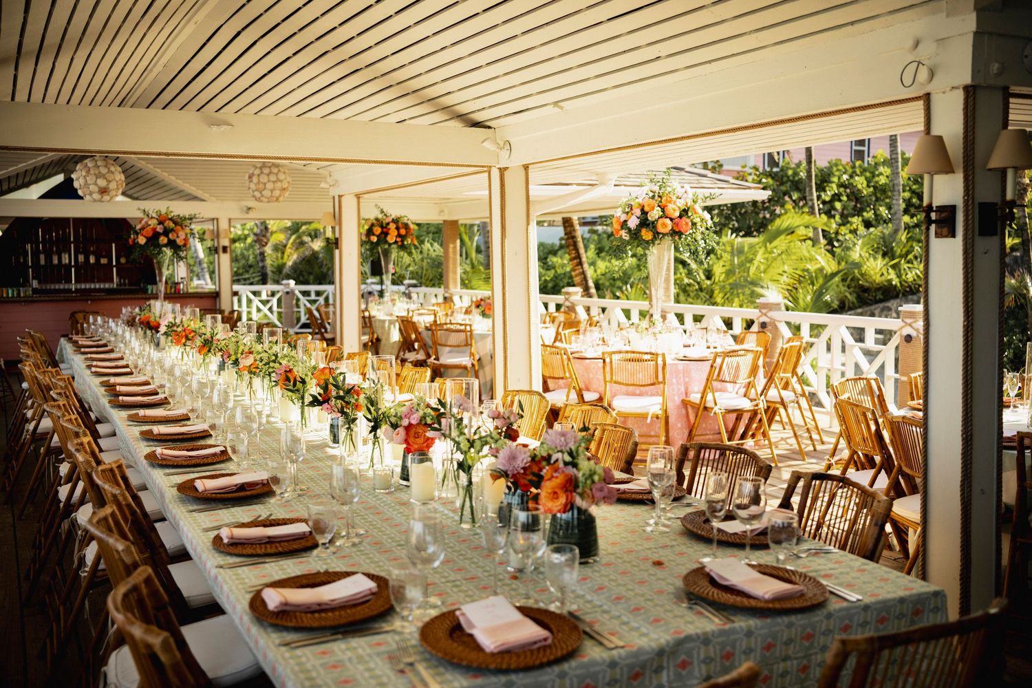Elegant outdoor wedding reception setup with long tables, wooden chairs and floral centerpieces under a covered patio.