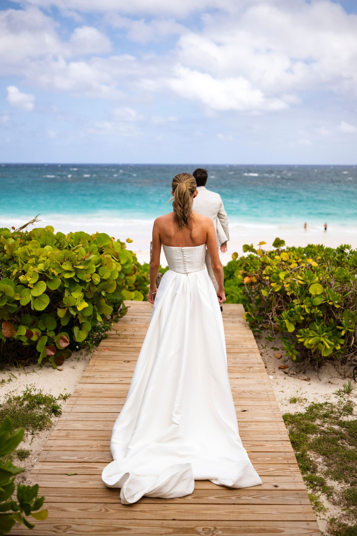 A couple in wedding attire walks along a wooden boardwalk surrounded by tropical foliage on a beach.