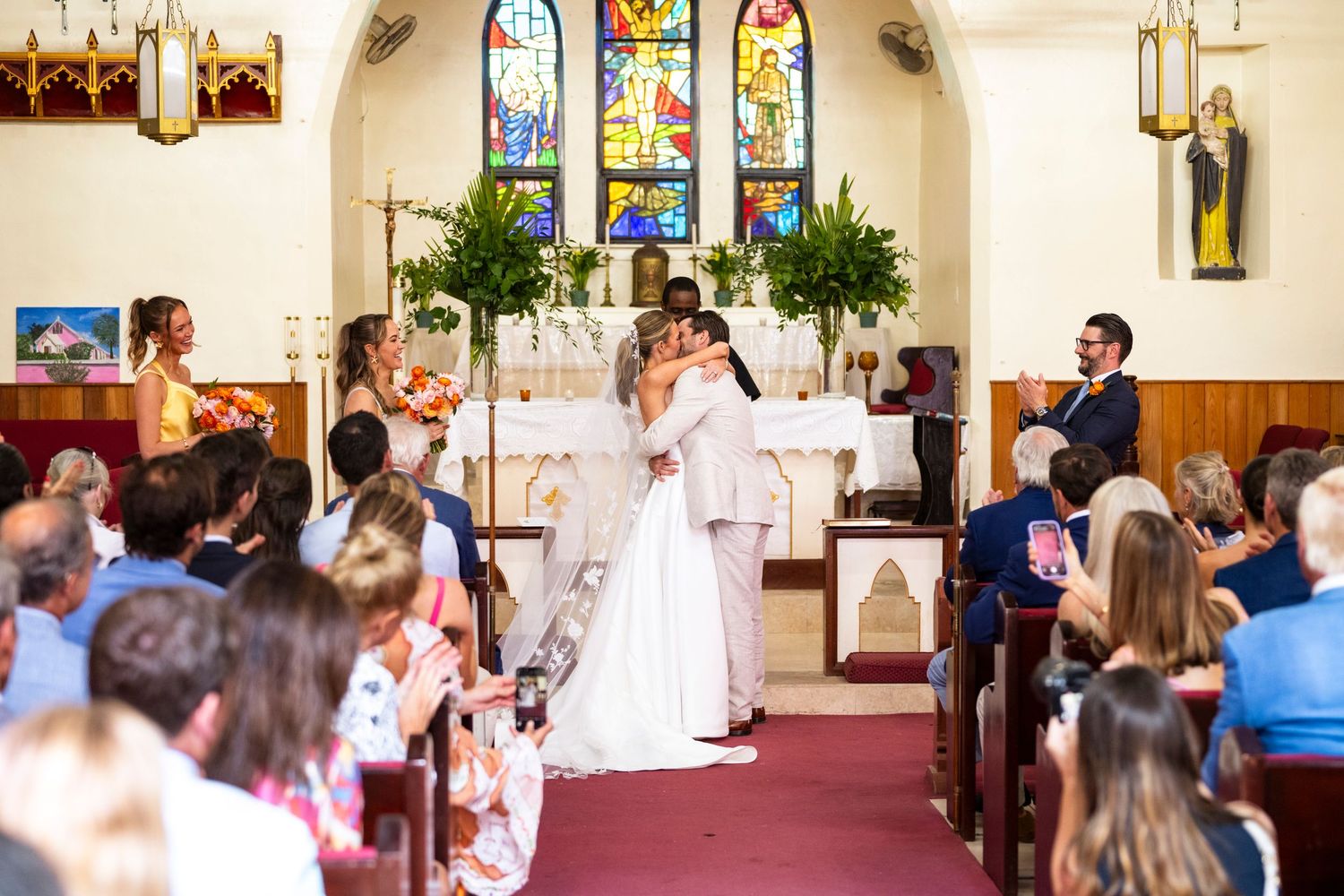 Wedding ceremony takes place in a church with stained glass windows and guests watching from wooden pews.