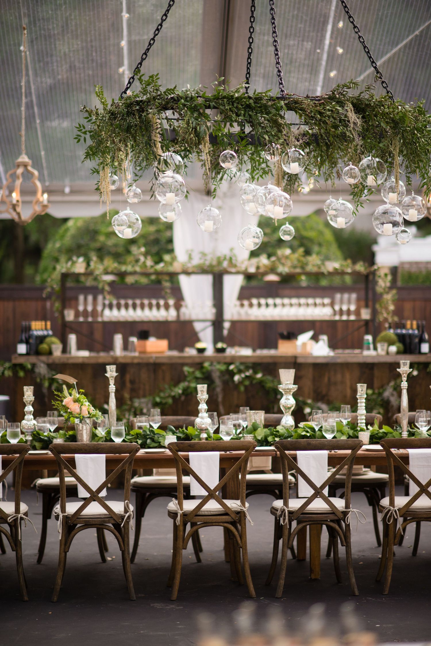 Rustic wooden cross-back chairs line long reception tables decorated with hanging glass orbs and greenery at a wedding venue.