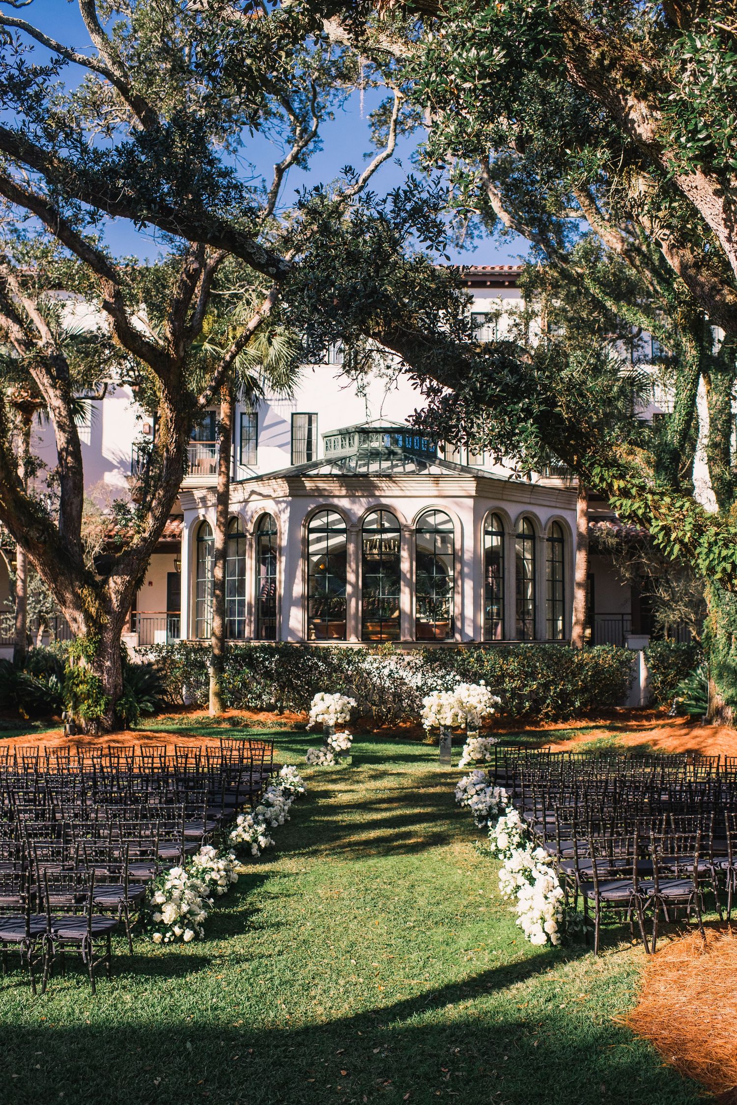 Rows of black chairs arranged on a lawn for an outdoor wedding ceremony with white floral arrangements and a mansion backdrop.