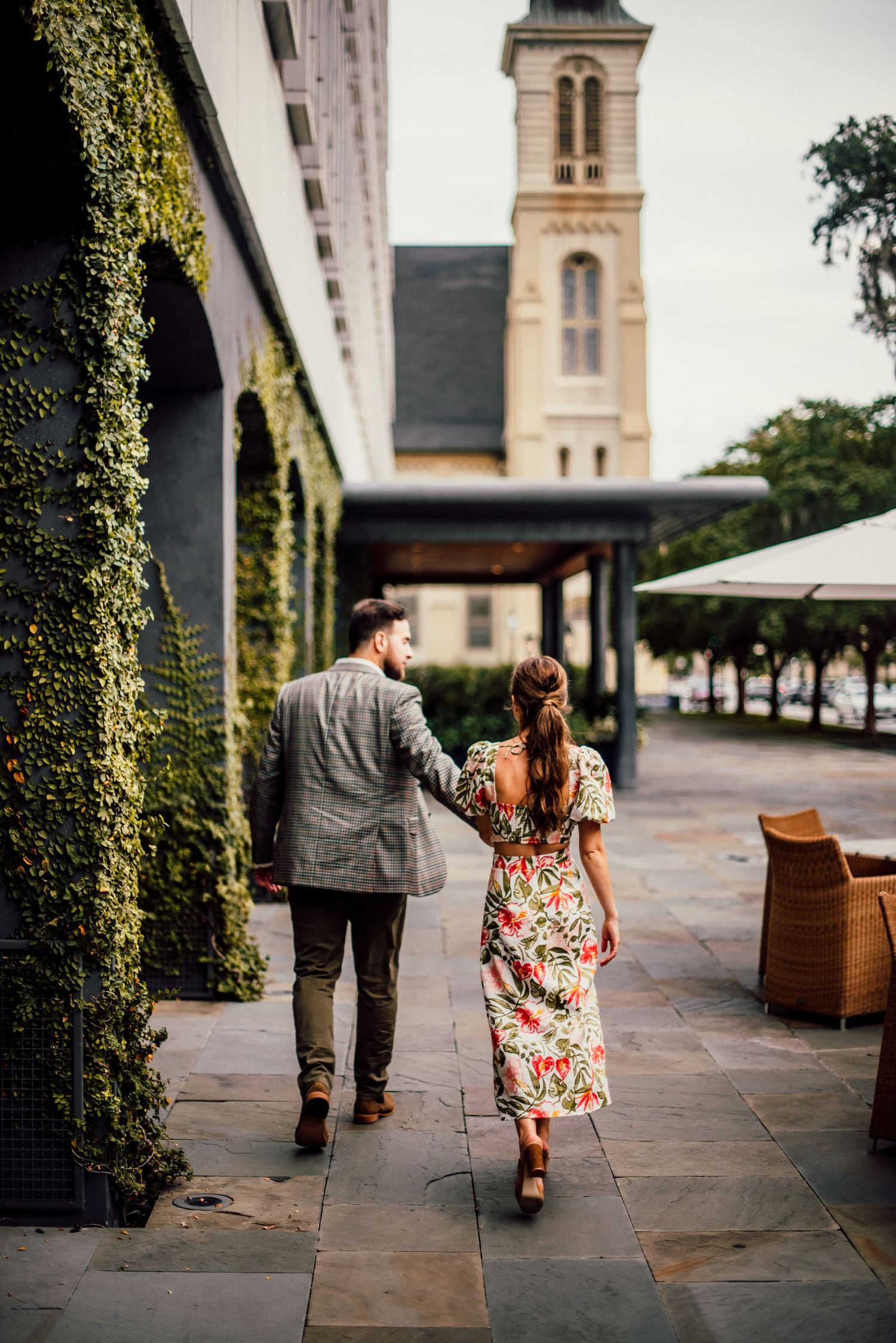 Two people walk together on a stone patio near a church building with ivy-covered walls in the background.