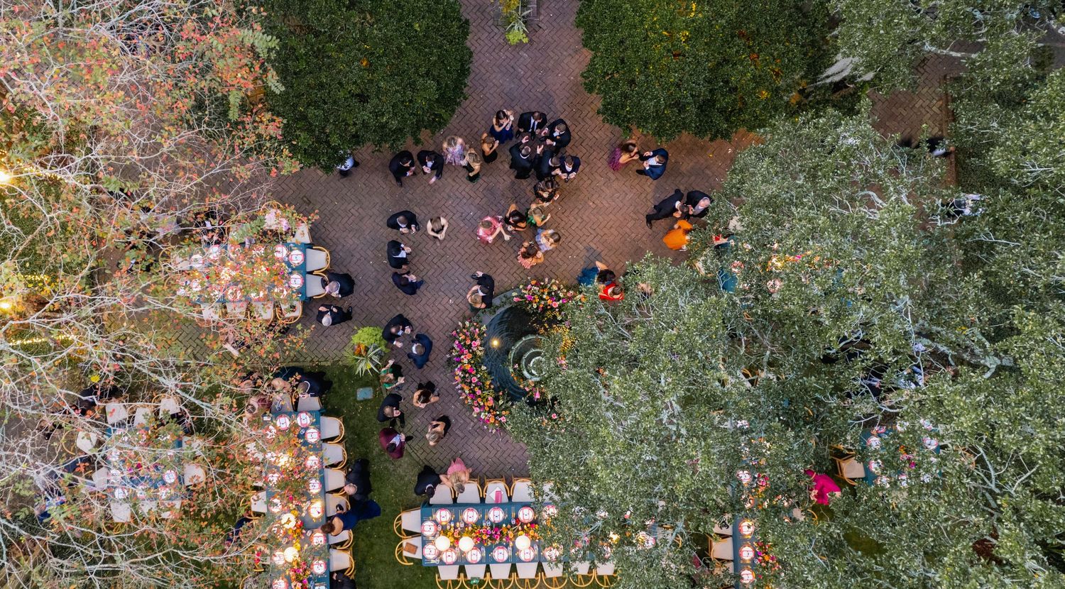 Aerial view of a large outdoor wedding ceremony with white chairs arranged in rows under green trees.