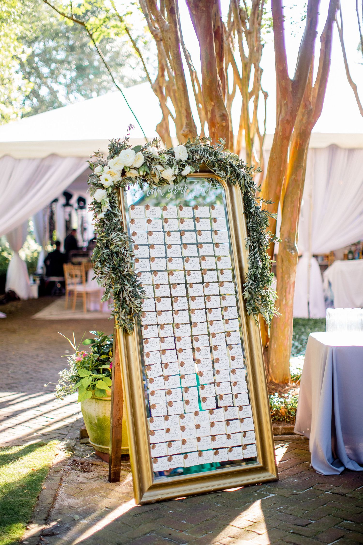 Elegant wedding seating chart display with greenery arch and white frames arranged in grid pattern under outdoor tent.