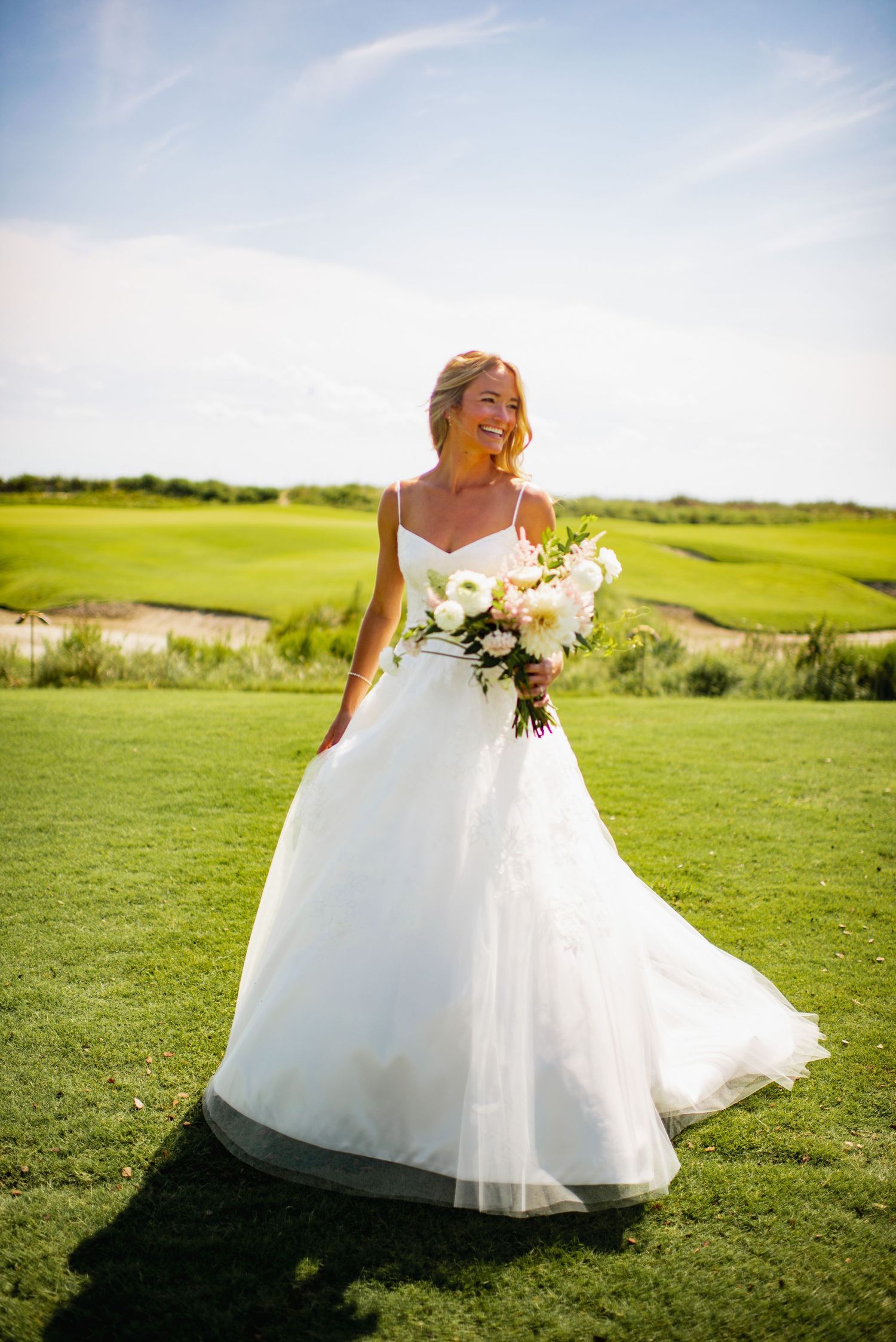A bride in a flowing white gown holds a bouquet while standing on a lush green golf course under blue skies.