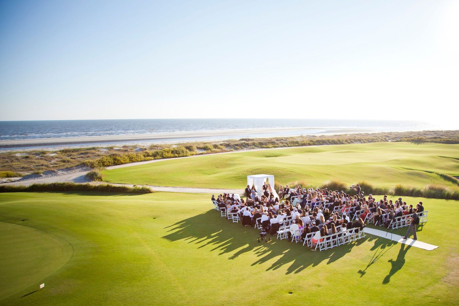 Large wedding party gathers for group photo on scenic golf course overlooking the ocean on a sunny day.