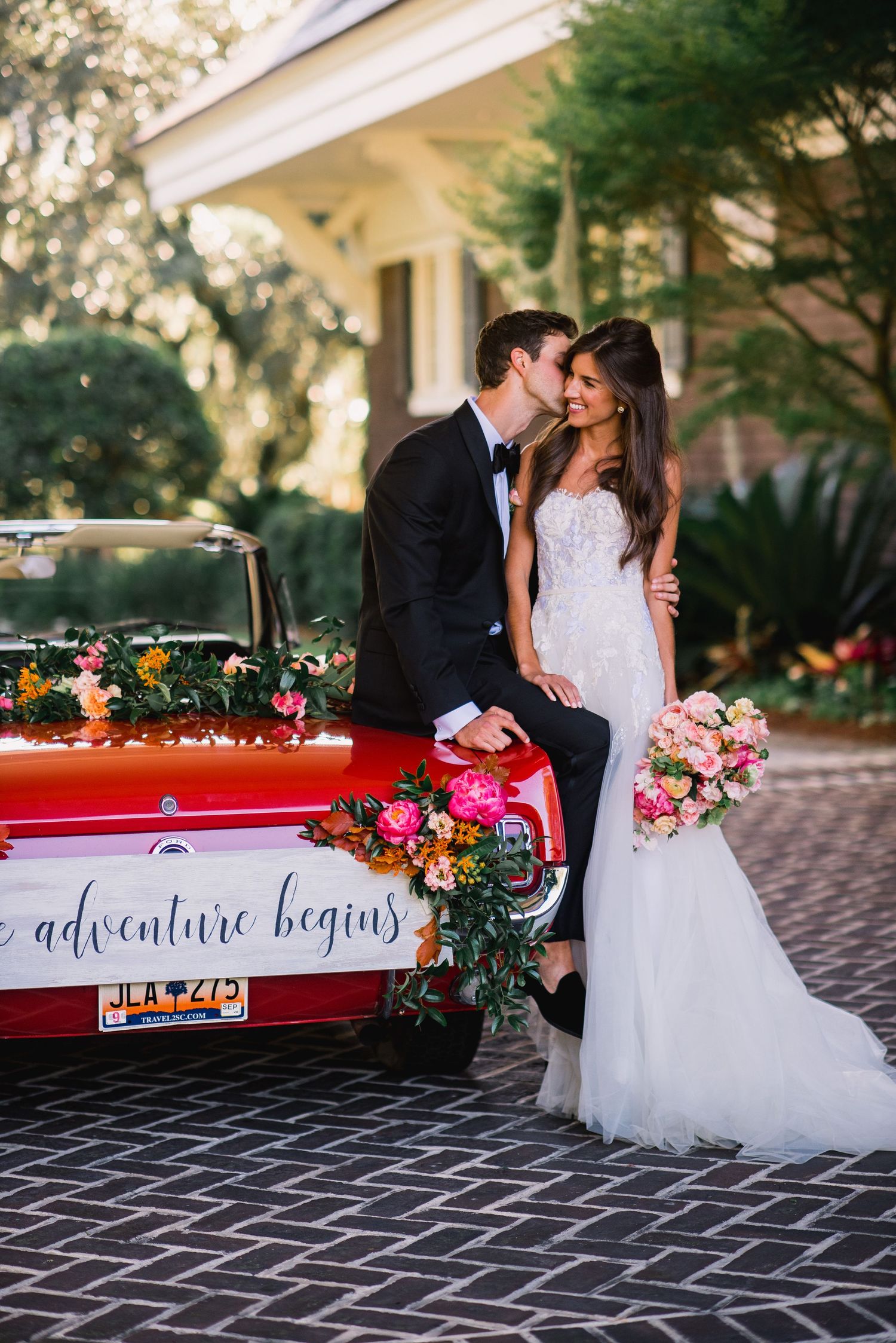 A romantic wedding couple poses with a vintage red car decorated with flowers and 'adventure begins' text on cobblestone.