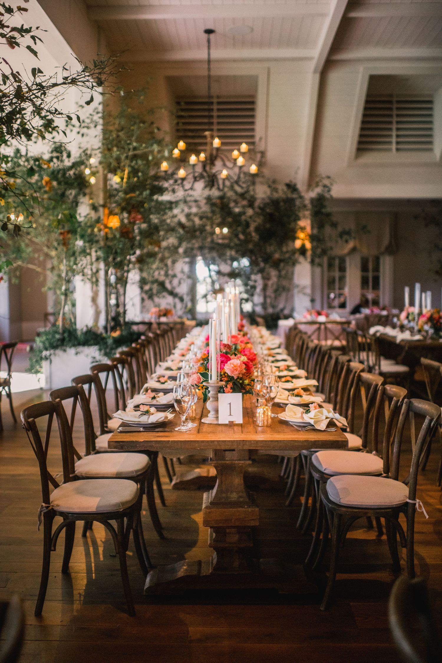 Rustic wedding reception venue with long wooden tables, crossback chairs, and hanging greenery in warm lighting.