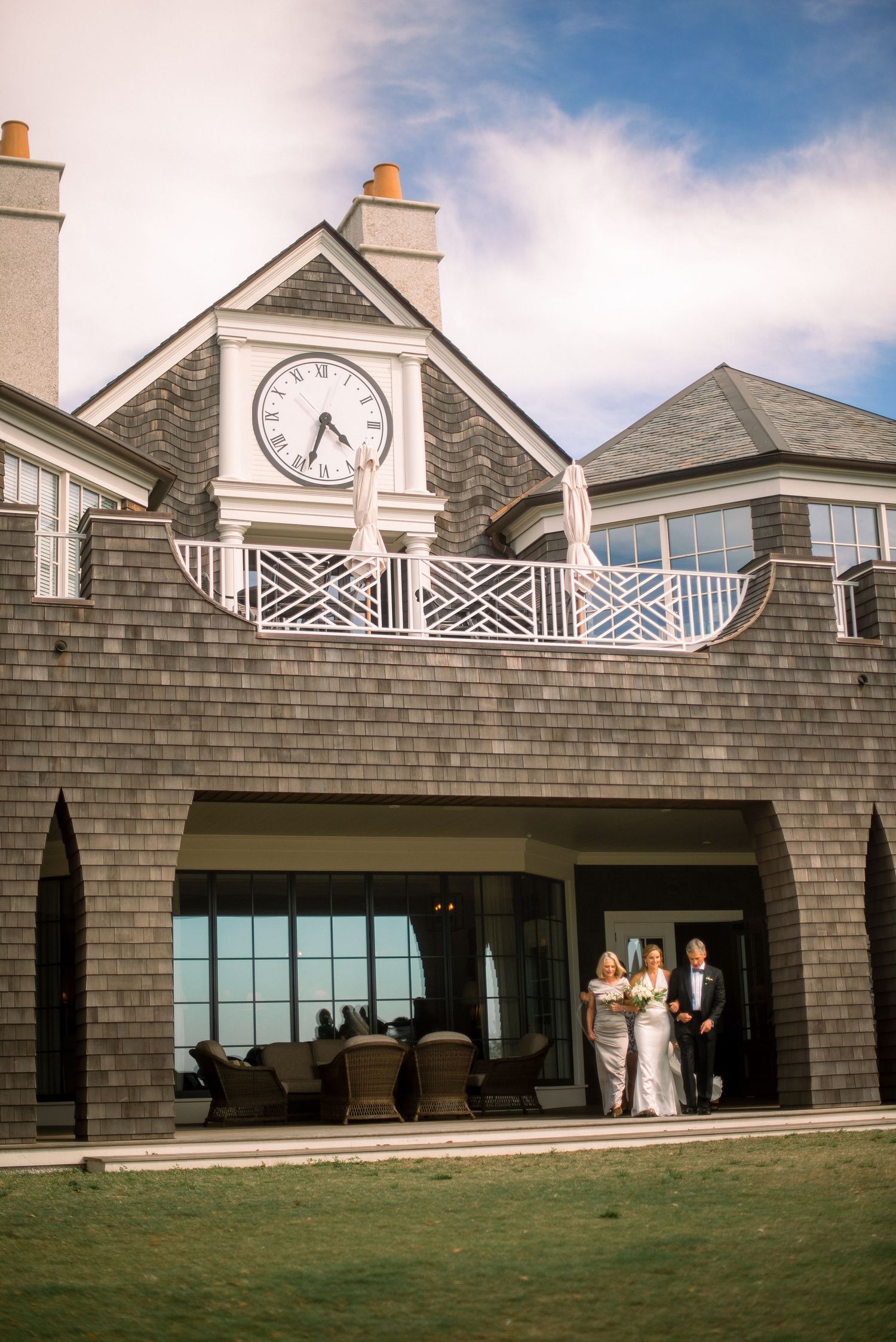 Elegant coastal mansion with large clock feature and white railings overlooking the ocean on a sunny day.