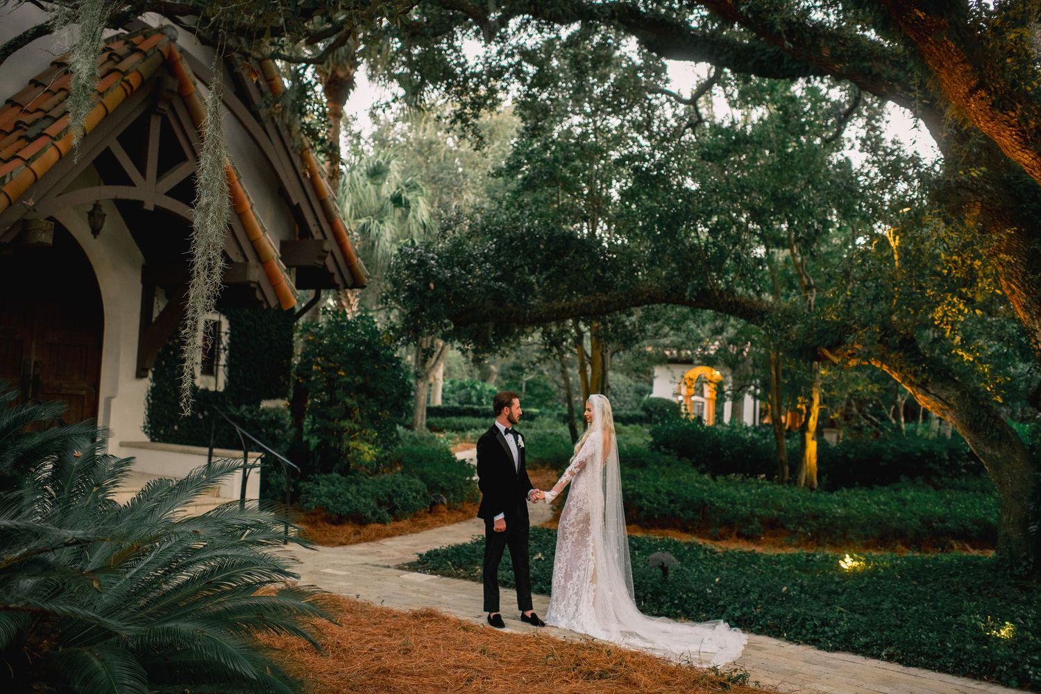 Wedding couple walks under majestic oak trees at sunset in a romantic garden setting.