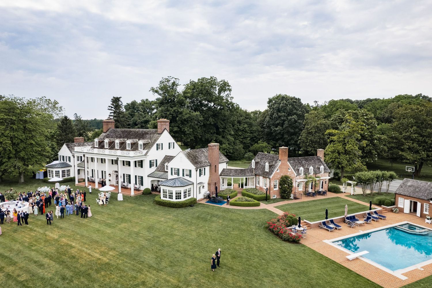 Aerial view of a grand white colonial estate with manicured lawn, swimming pool and guests gathered for an outdoor event.