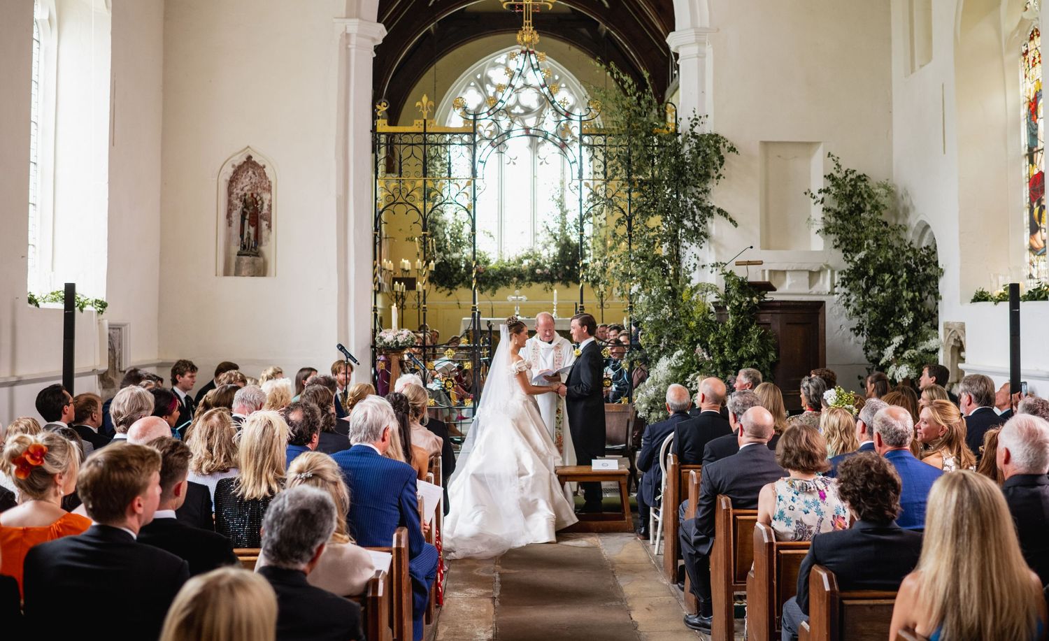 Wedding ceremony takes place in a bright white church sanctuary with guests seated in wooden pews.