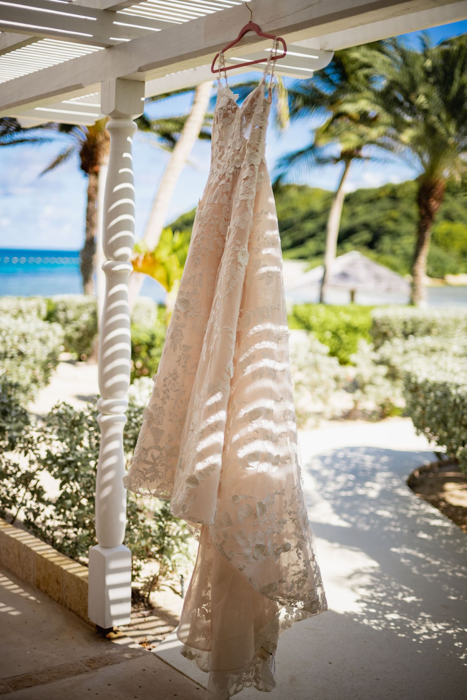 White wedding dress hanging from a tropical beach pergola with palm trees and ocean in background.